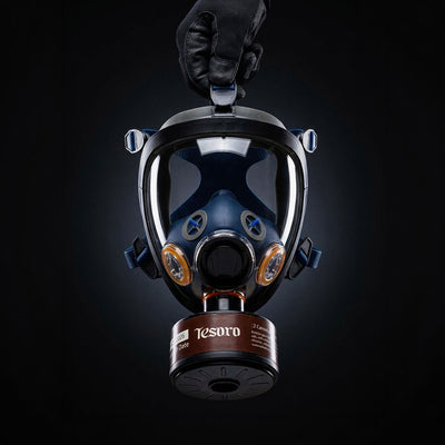 Gas mask with 'Tesoro' branding held by a gloved hand against a black background