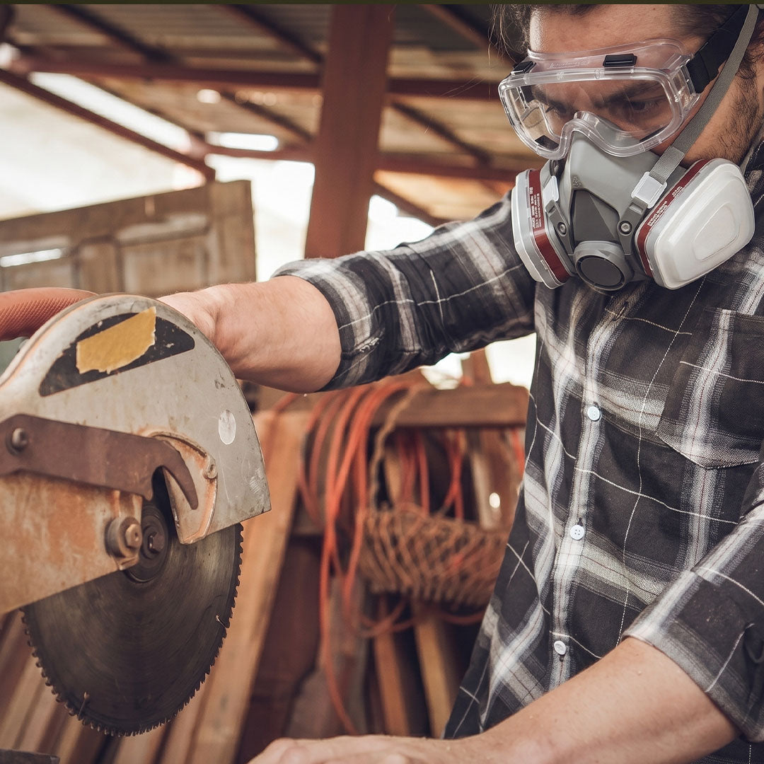 Person using a circular saw with safety goggles and a mask in a workshop setting.